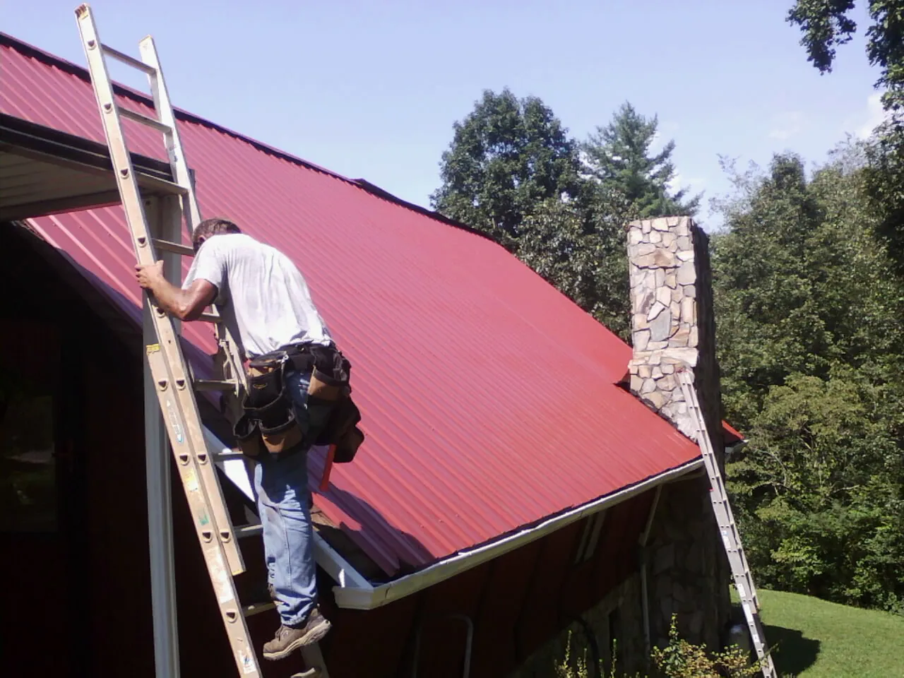 Roofer on a ladder beside a red metal roof and stone chimney — Soffit & Fascia Repair by Roofing Brothers in Brush Valley
